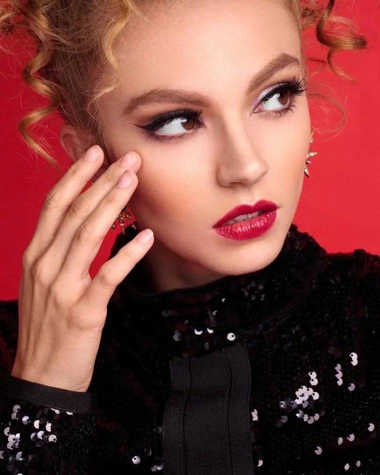 Model with a curled updo, winged liner and red lips against a red backdrop
